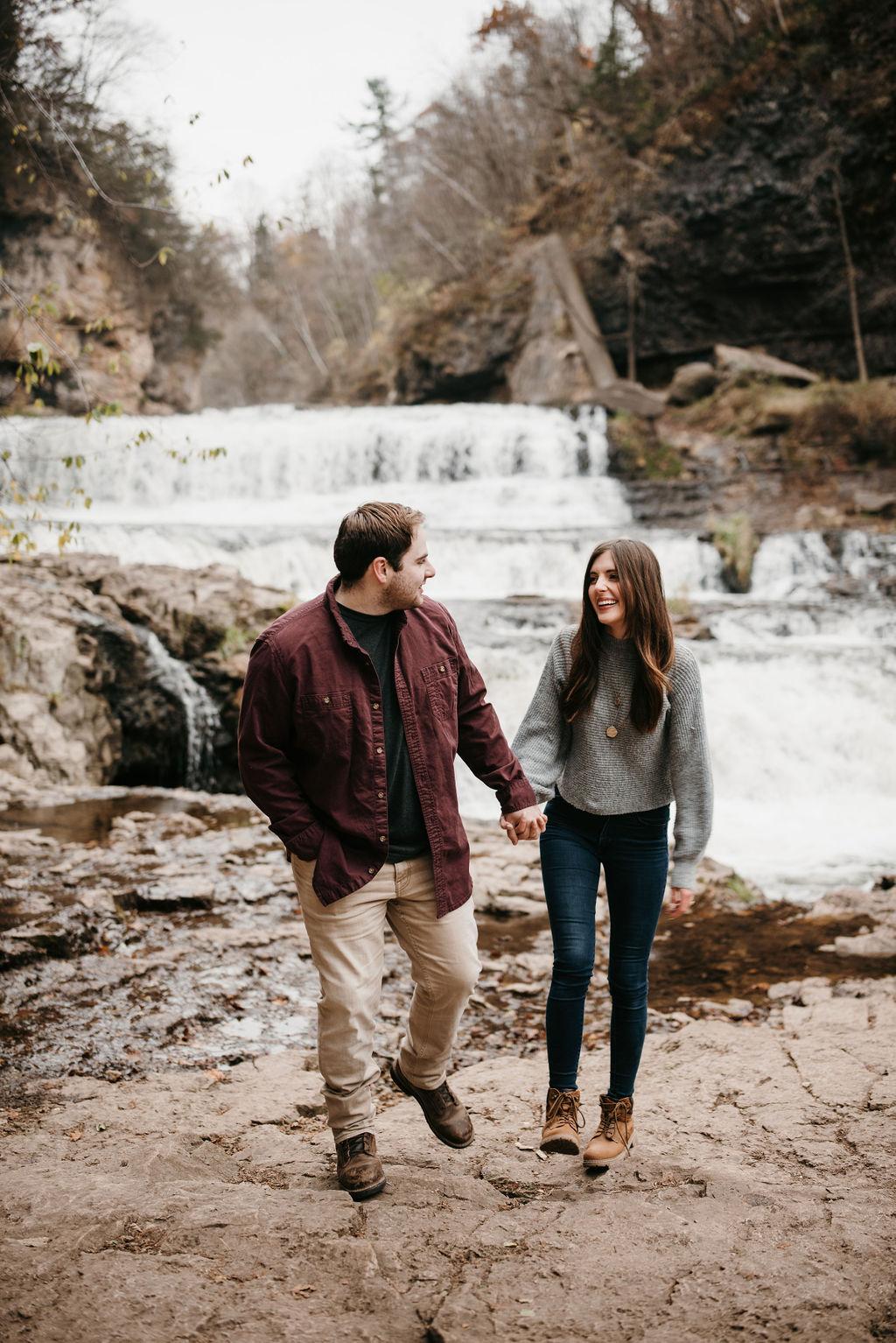 Morgan and Garrett are holding hands and walking in front of a waterfall and cliffs.
